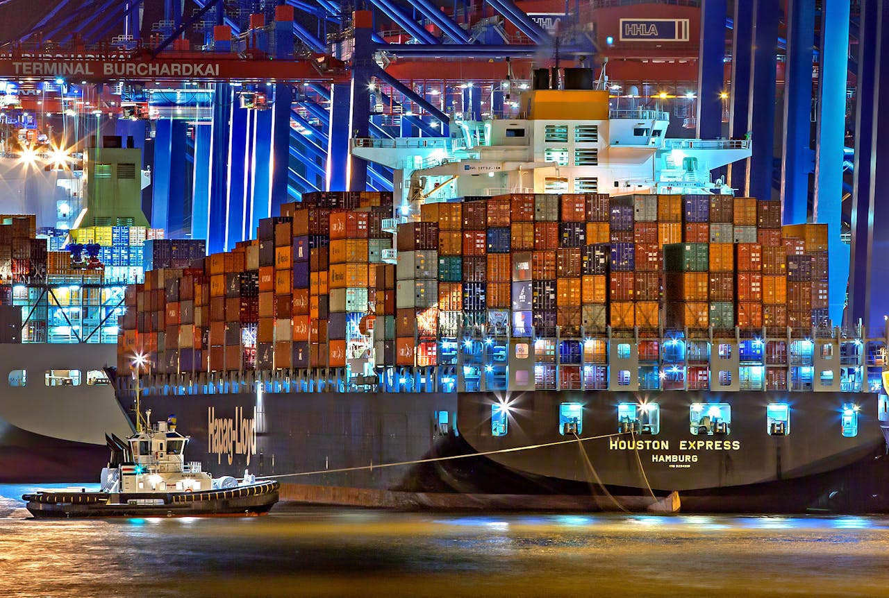 services-05 A brightly lit cargo ship at Hamburg harbor with stacked containers and a tugboat.